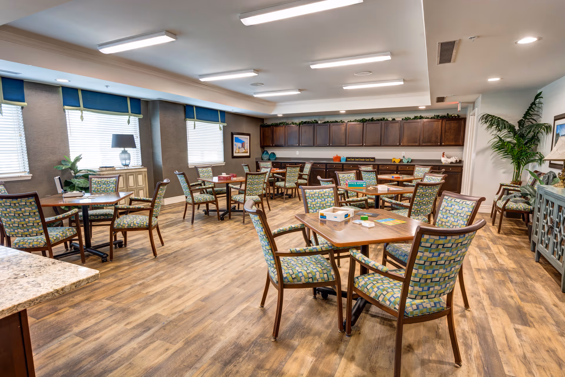 A spacious, well-lit common room with multiple wooden tables and chairs featuring patterned upholstery. The room has large windows with blue valances, wooden flooring, and a long cabinet with decorative items along the back wall. There are plants and framed pictures adding to the decor.