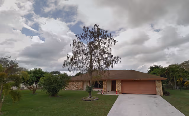 Single-story house with a stone facade and a brown roof, featuring a two-car garage with a brown door. A large tree with sparse leaves stands in the front yard, which is covered with green grass and surrounded by other smaller trees and shrubs. The sky above is cloudy with patches of blue.