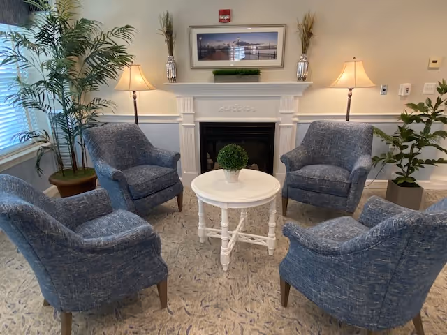 A cozy seating area with four blue upholstered armchairs arranged around a small white round table with a potted plant on top. Behind the chairs is a white fireplace mantel with decorative molding, two table lamps with beige shades on either side, and a framed picture hanging above. There are also two tall plants in pots on either side of the fireplace.