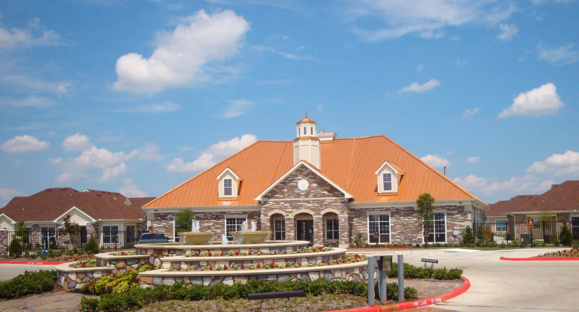 Front exterior view of a senior living facility building with a stone facade and a large orange metal roof under a blue sky with scattered clouds. In front of the building is a tiered stone fountain surrounded by landscaping and a circular driveway with red curbs.