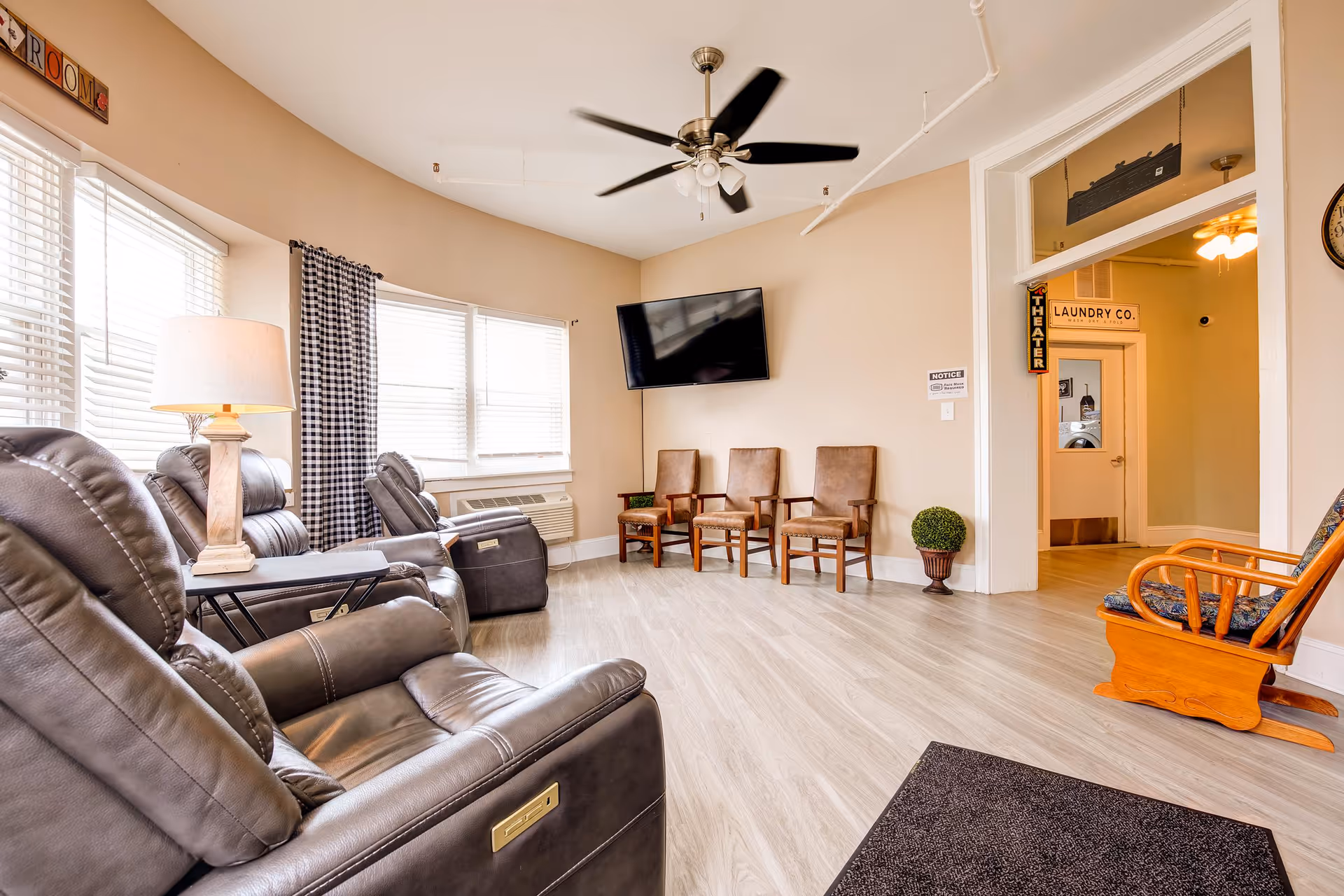 A bright and spacious senior living common area with several leather recliners, wooden chairs, a rocking chair, a ceiling fan, a wall-mounted flat screen TV, and large windows with blinds and checkered curtains. There is a lamp on a side table and a small potted plant near the doorway, which leads to a hallway with signs for 'Laundry Co.' and 'Theater'.