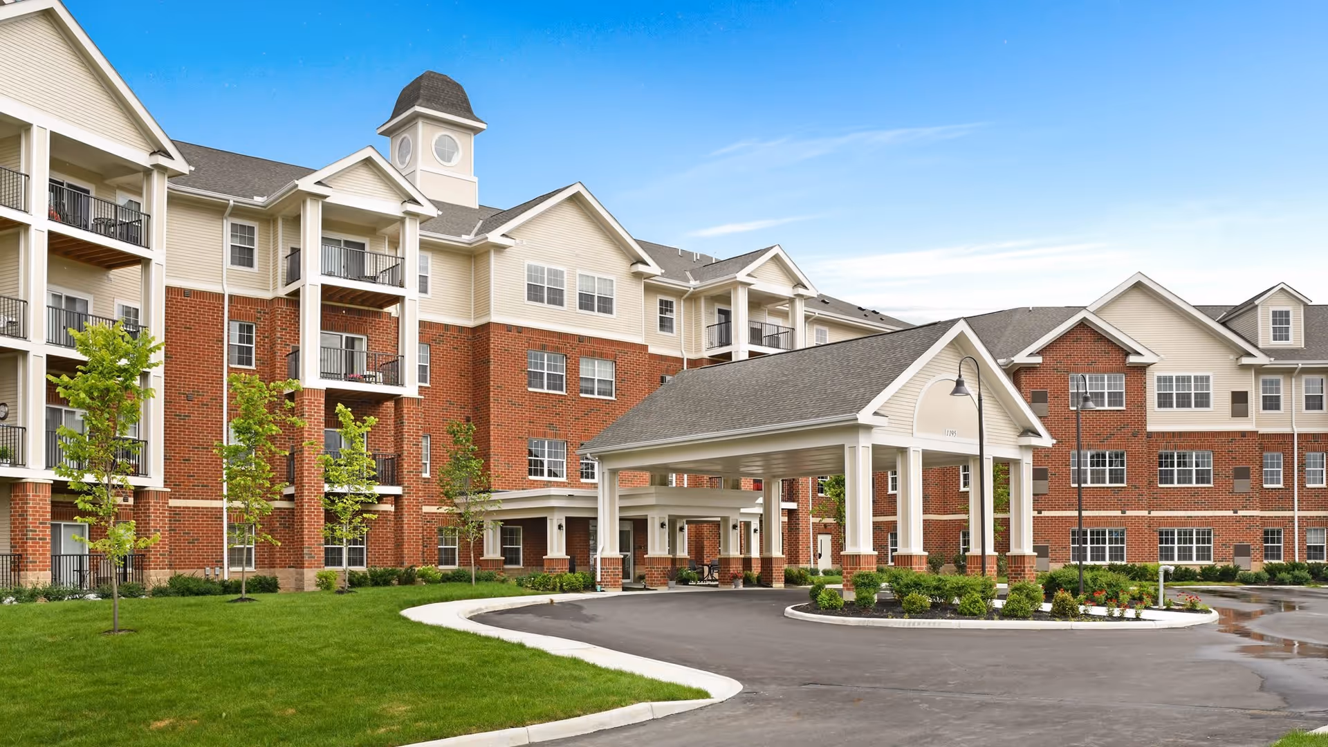 Exterior view of a multi-story senior living facility with red brick and beige siding, featuring balconies, a covered entrance driveway, green lawn, and young trees under a clear blue sky.