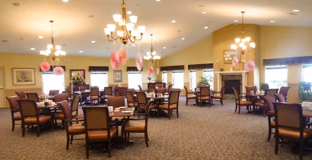 A spacious dining room with multiple wooden tables and chairs arranged neatly. The room is well-lit with chandeliers and large windows allowing natural light. Pink paper decorations hang from the ceiling. There is a fireplace on the far wall with plants and framed artwork around the room.