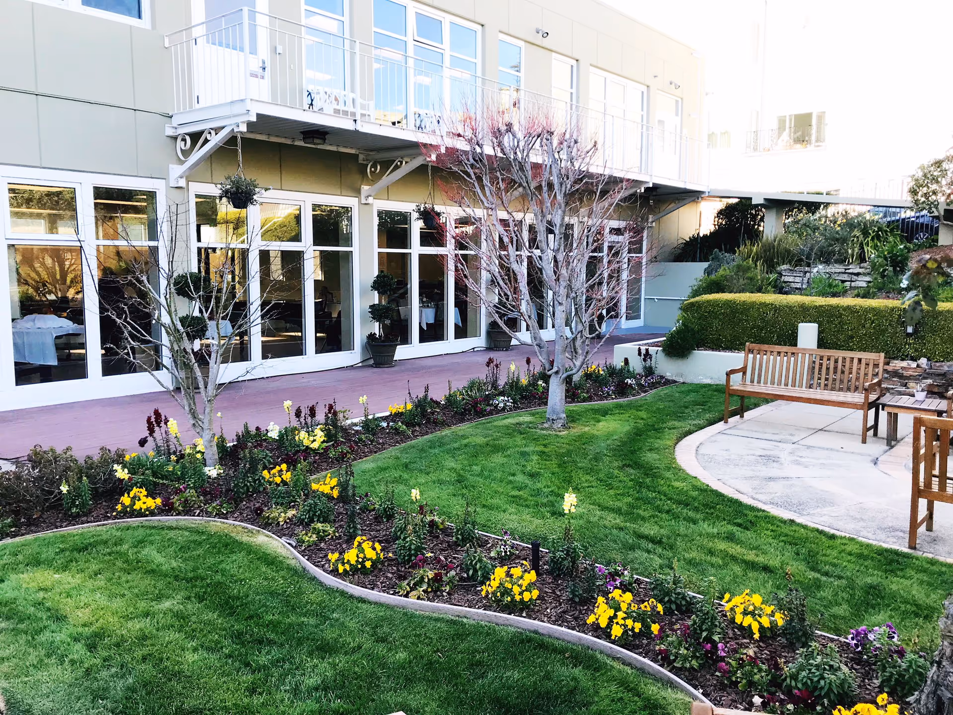 Outdoor garden area at Sagebrook Senior Living featuring a well-maintained lawn, flower beds with yellow and purple flowers, leafless trees, and wooden benches on a paved circular patio. The building with large windows and a balcony is visible in the background.