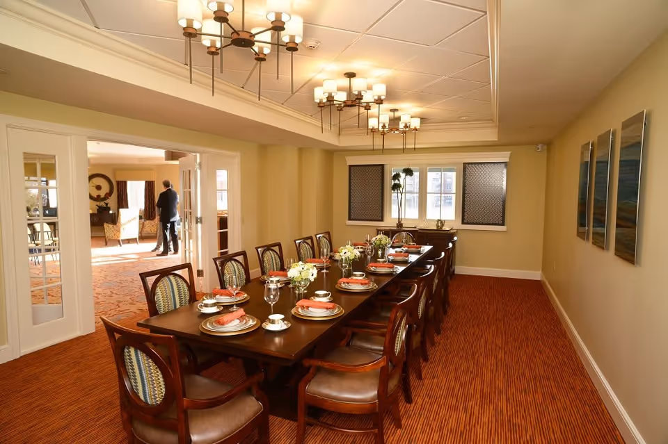 A well-lit dining room with a long wooden table set for twelve people. Each place setting includes a plate, cup, saucer, glass, and a folded orange napkin. The room has beige walls, three framed artworks on the right wall, and two windows at the far end with decorative coverings. Two chandeliers hang from the ceiling. Through open double doors, another room with seating and a person standing is visible.