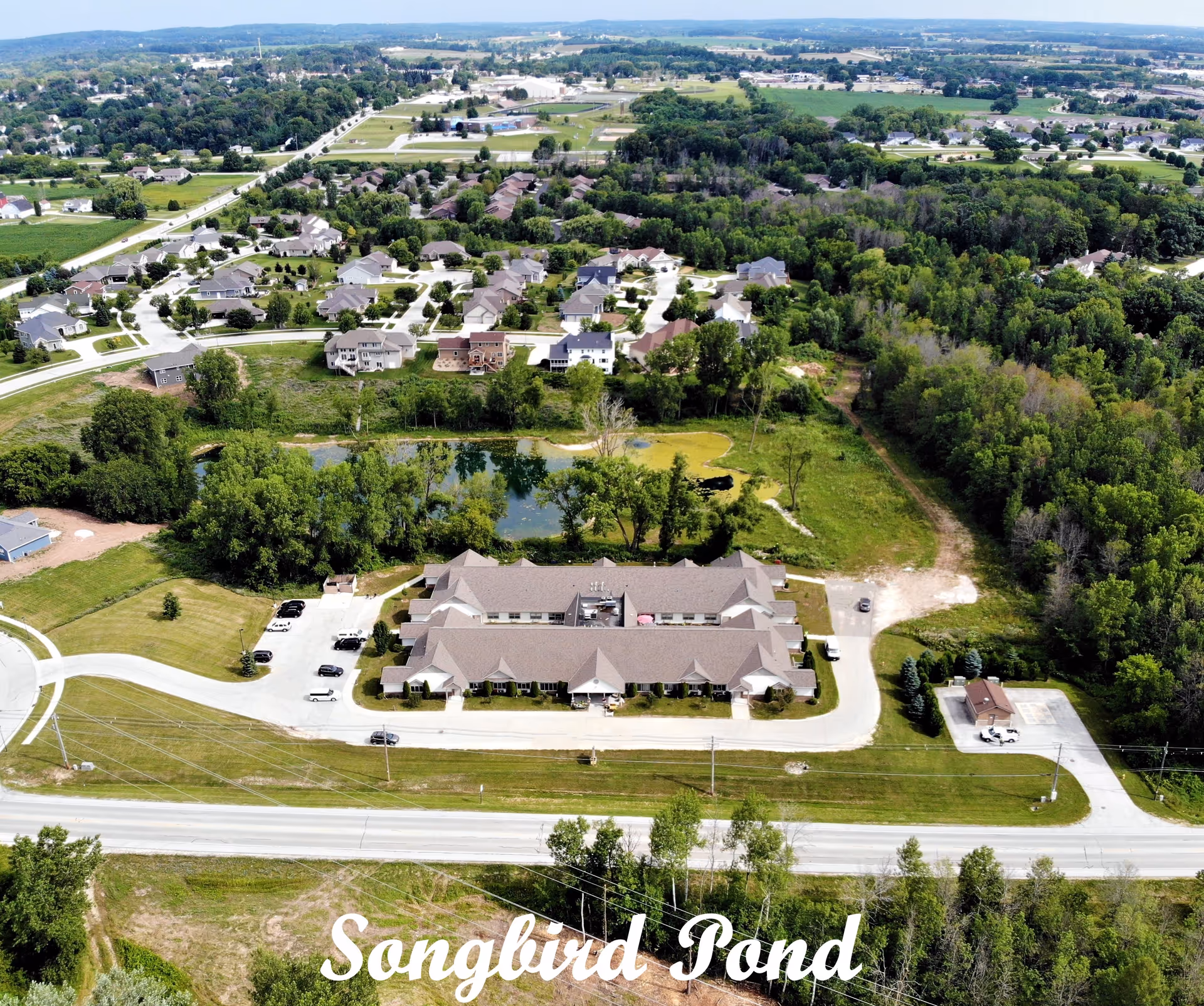 Aerial view of Songbird Pond Assisted Living facility surrounded by greenery, a pond, and nearby residential houses. The building is a single-story structure with a parking area and a road in front.