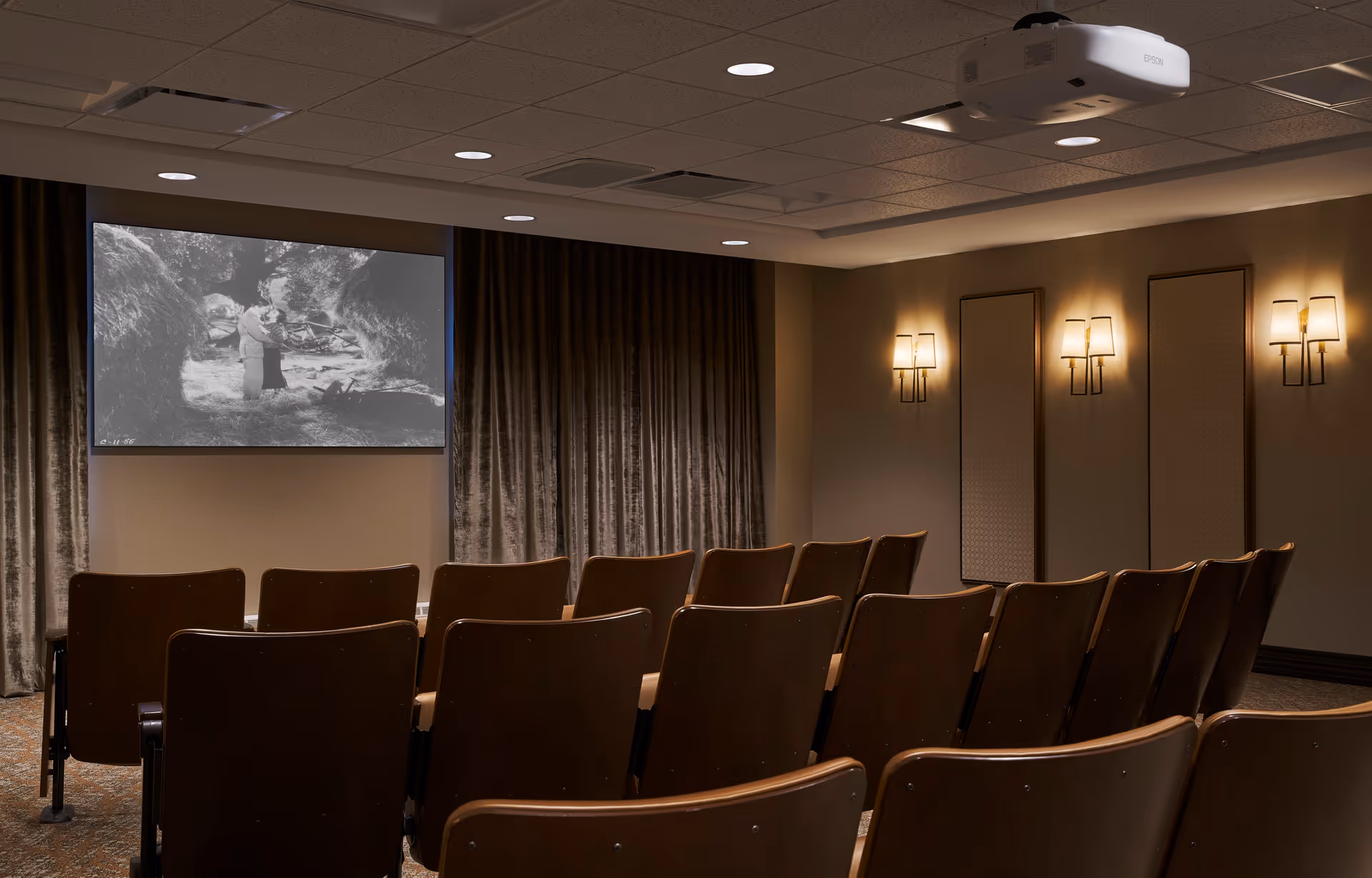 A small theater room with rows of brown chairs facing a large screen showing a black and white movie. The room has beige walls, wall-mounted lights, and a ceiling-mounted Epson projector.