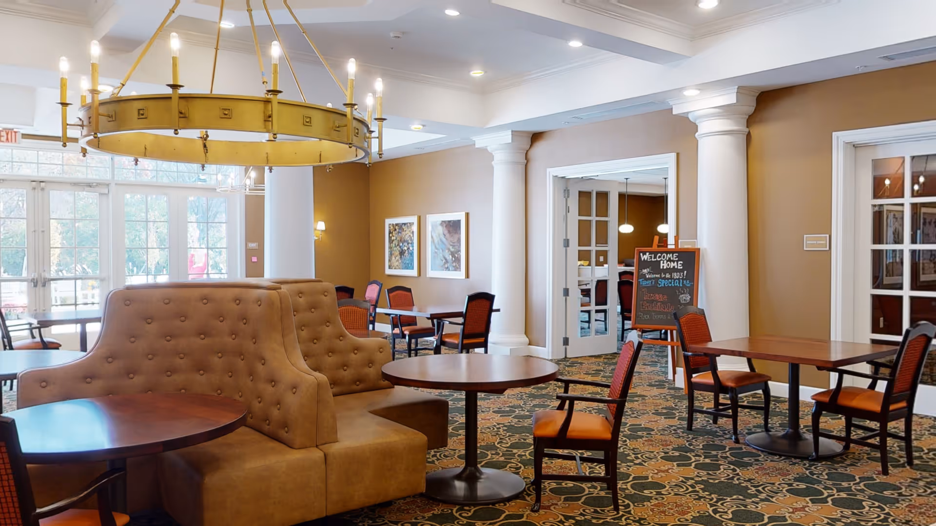 A spacious and well-lit common area in a senior living facility with round wooden tables and cushioned chairs arranged for socializing or dining. The room features a large, circular chandelier with candle-like lights hanging from the ceiling, beige walls with white columns, framed artwork, and a patterned carpet. A chalkboard sign near a doorway reads 'Welcome Home' with additional text about specials.