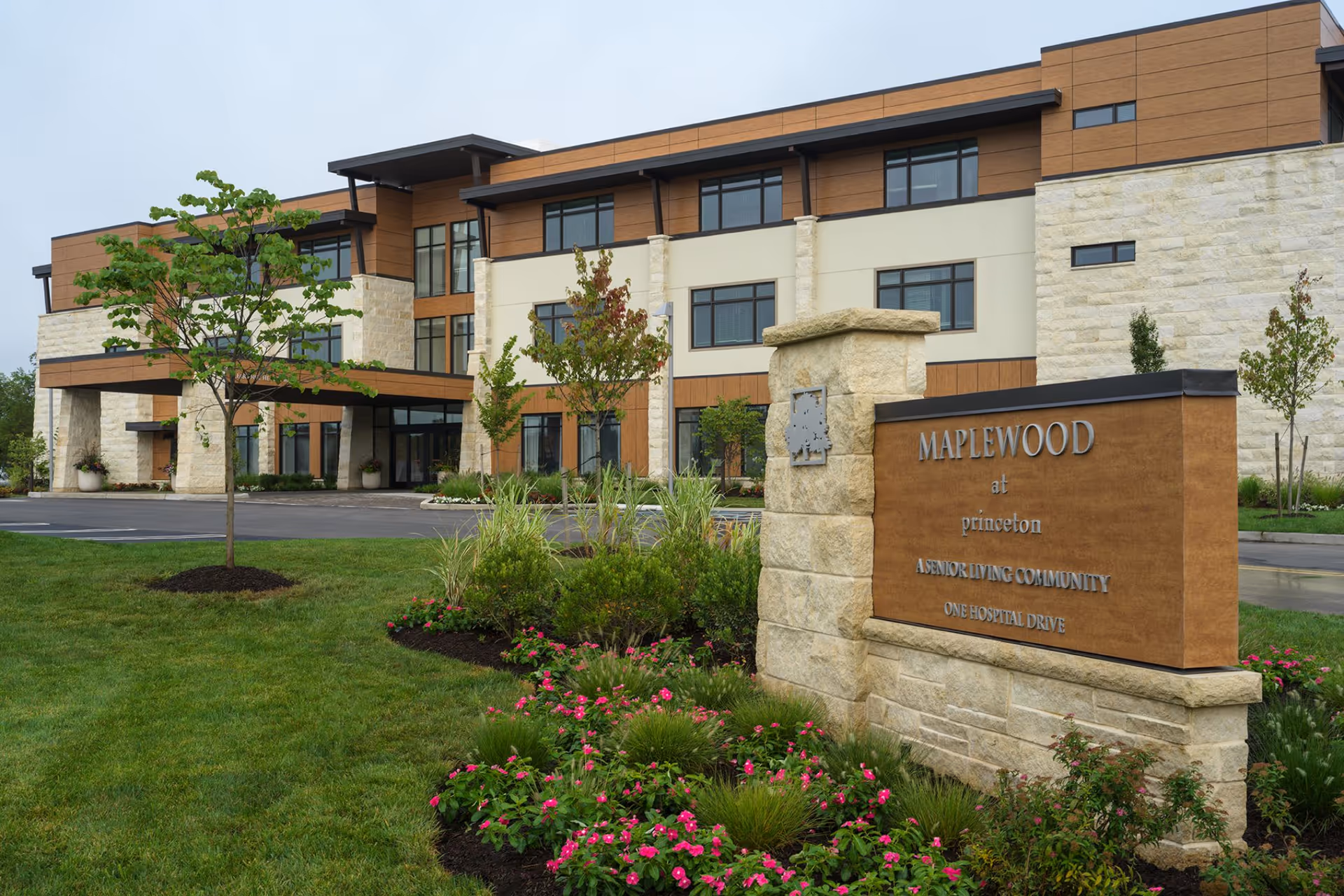 Exterior view of Maplewood at Princeton, a senior living community building with a modern design featuring stone and wood paneling. The foreground shows a landscaped garden with green grass, small trees, and pink flowers, along with a stone sign displaying the facility's name and address.