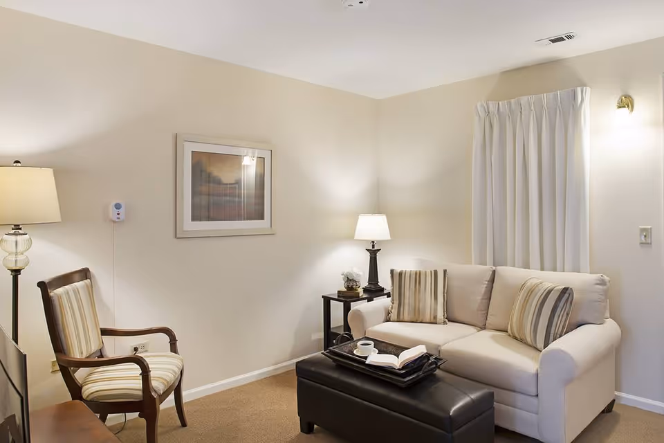 A cozy living room with a beige sofa adorned with striped cushions, a black ottoman serving as a coffee table with an open book and a cup on a tray, a wooden armchair with striped upholstery, a floor lamp, a side table with a lamp and decorative items, a framed picture on the wall, and white curtains covering a window.