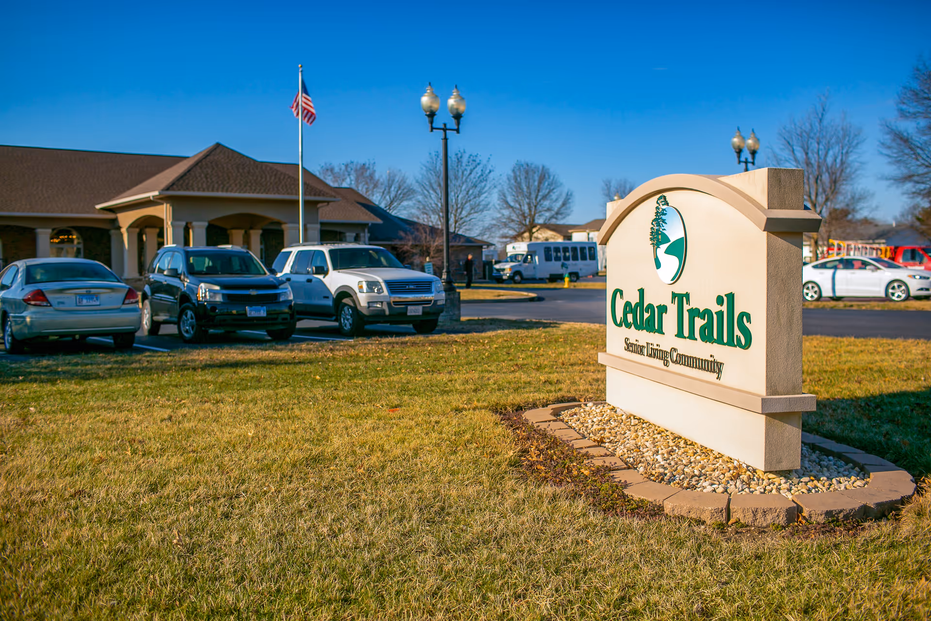 Exterior view of Cedar Trails Senior Living Community sign on a grassy area with a building, parked cars, and an American flag in the background under a clear blue sky.