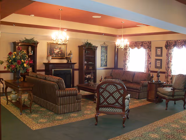 A cozy living room area with two striped sofas, two patterned armchairs, a wooden coffee table, and a fireplace with decorative items on the mantel. The room features floral curtains on the windows, two chandeliers hanging from the ceiling, and a large floral arrangement on a wooden side table.
