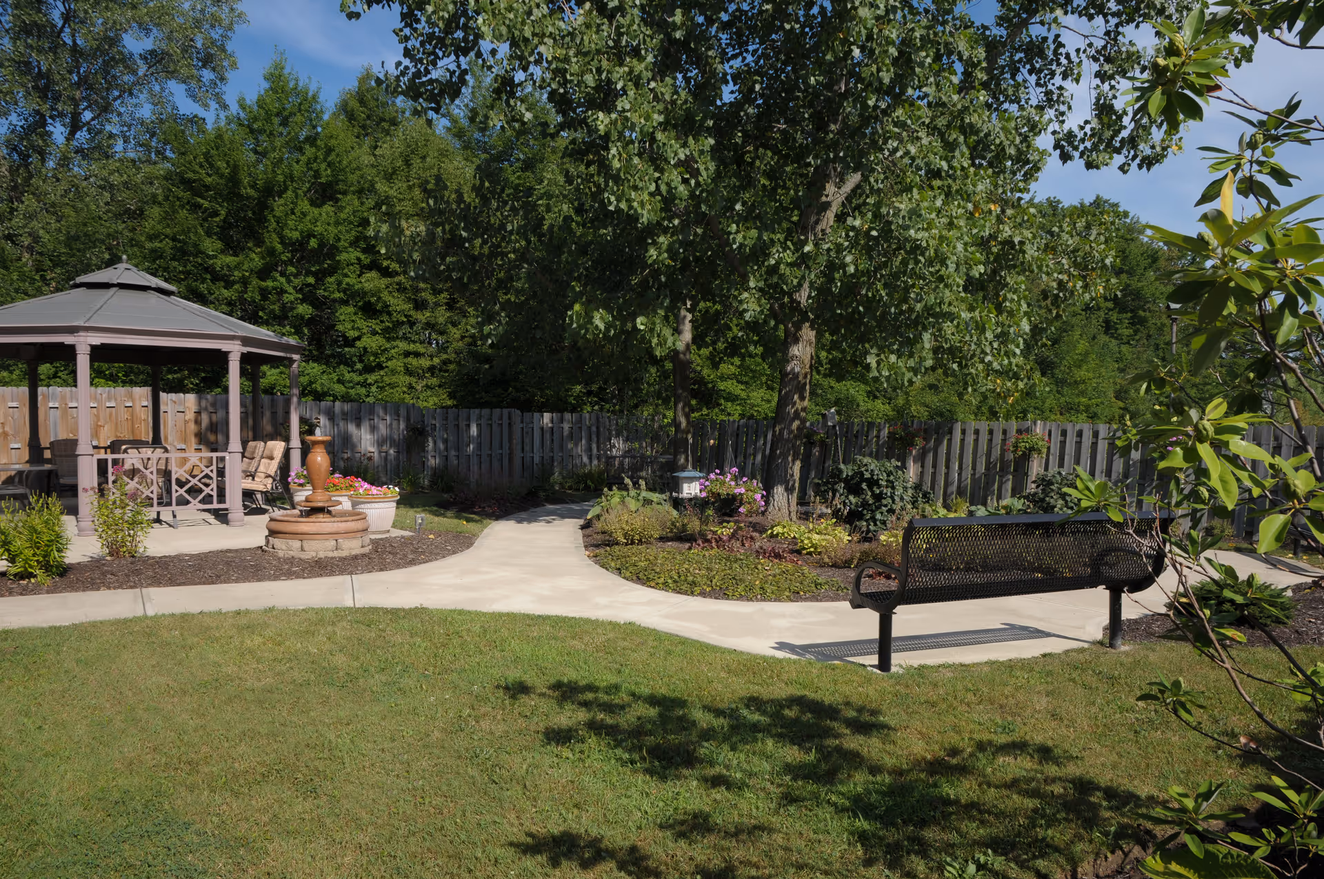 A peaceful outdoor garden area with a paved walkway, a black metal bench, a gazebo with chairs, a small fountain, and various plants and trees surrounded by a wooden fence under a clear blue sky.
