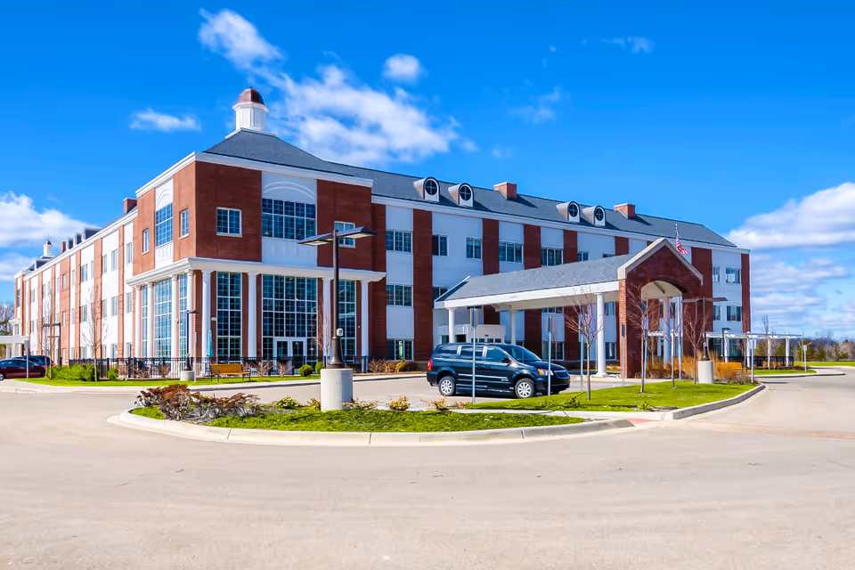 Exterior view of a large, three-story senior living facility building with red brick and white paneling under a blue sky with scattered clouds. There is a covered entrance with a vehicle parked underneath and a landscaped roundabout in front.