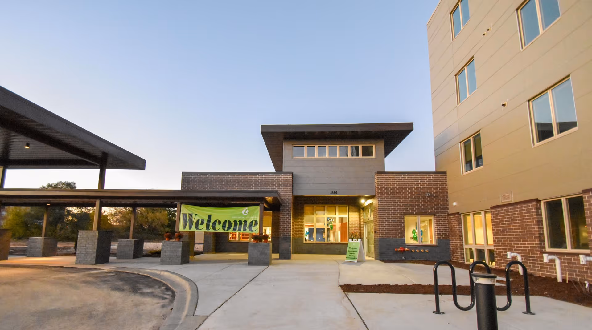 Exterior view of The Sonnet at Tennyson Assisted Living & Memory Care building entrance during dusk, featuring a covered drop-off area with a green 'Welcome' banner and well-lit windows.