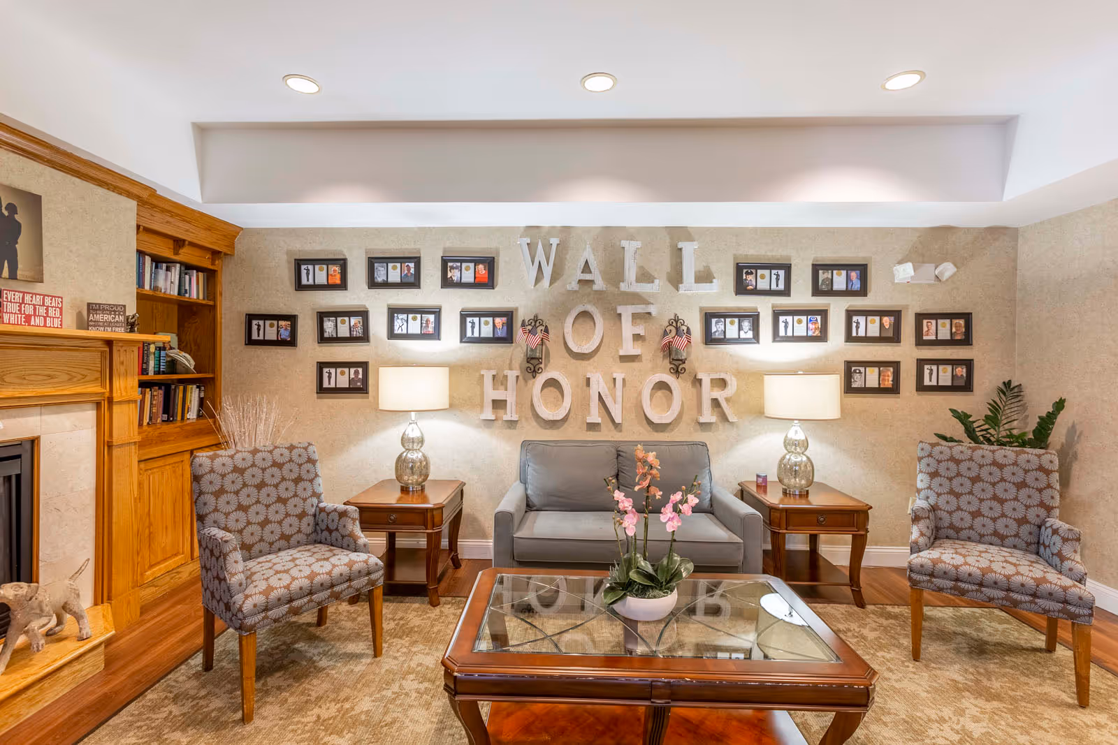 A cozy living room area with a gray loveseat centered between two wooden side tables with lamps. Two patterned armchairs flank the loveseat. A wooden coffee table with a glass top and a flower arrangement sits in front. The wall behind the loveseat displays the words 'WALL OF HONOR' with framed photos arranged around the text. To the left, there is a wooden bookshelf and a fireplace with decorative items.