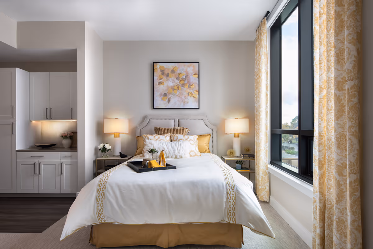 A bright and cozy bedroom featuring a neatly made bed with white and gold bedding, two bedside tables with lamps, a framed floral artwork above the headboard, and a large window with patterned yellow curtains letting in natural light. To the left, there is a small kitchenette area with white cabinets and under-cabinet lighting.