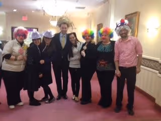 A group of seven people standing together in a hallway, some wearing colorful clown wigs and hats, smiling and posing for the photo.