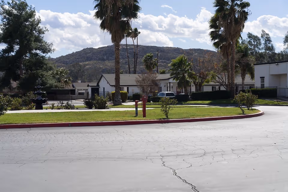 View of a senior living facility exterior with a paved driveway, green lawn, palm trees, and a white building in the background. Hills and a partly cloudy sky are visible behind the facility.