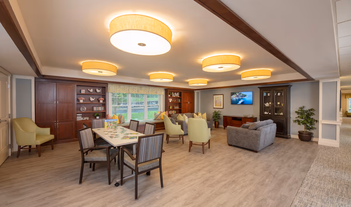 A spacious and well-lit common area in a senior living facility with a table and chairs in the foreground, comfortable armchairs and a sofa arranged around a wall-mounted TV, wooden cabinets and shelves with decorative items, large windows with a view of greenery outside, and modern ceiling lights.