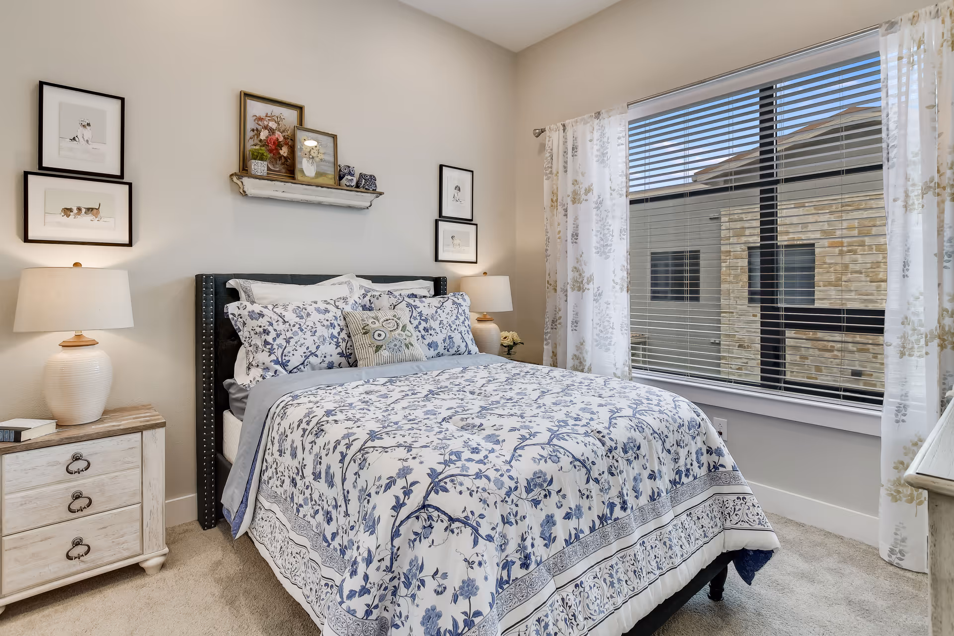 A cozy bedroom with a bed covered in white and blue floral bedding. There are two bedside tables with lamps on each side of the bed. Above the bed, there are framed pictures and a small shelf with decorative items. A large window with white and light gray patterned curtains lets in natural light, showing a view of a stone building outside.