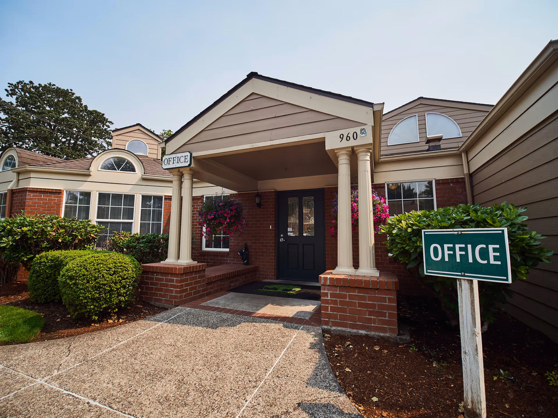 Exterior view of the entrance to the office at Faye Wright Senior Living, featuring a covered porch with columns, brick walls, a black door, hanging flower baskets, and a green sign labeled 'OFFICE' near trimmed bushes.