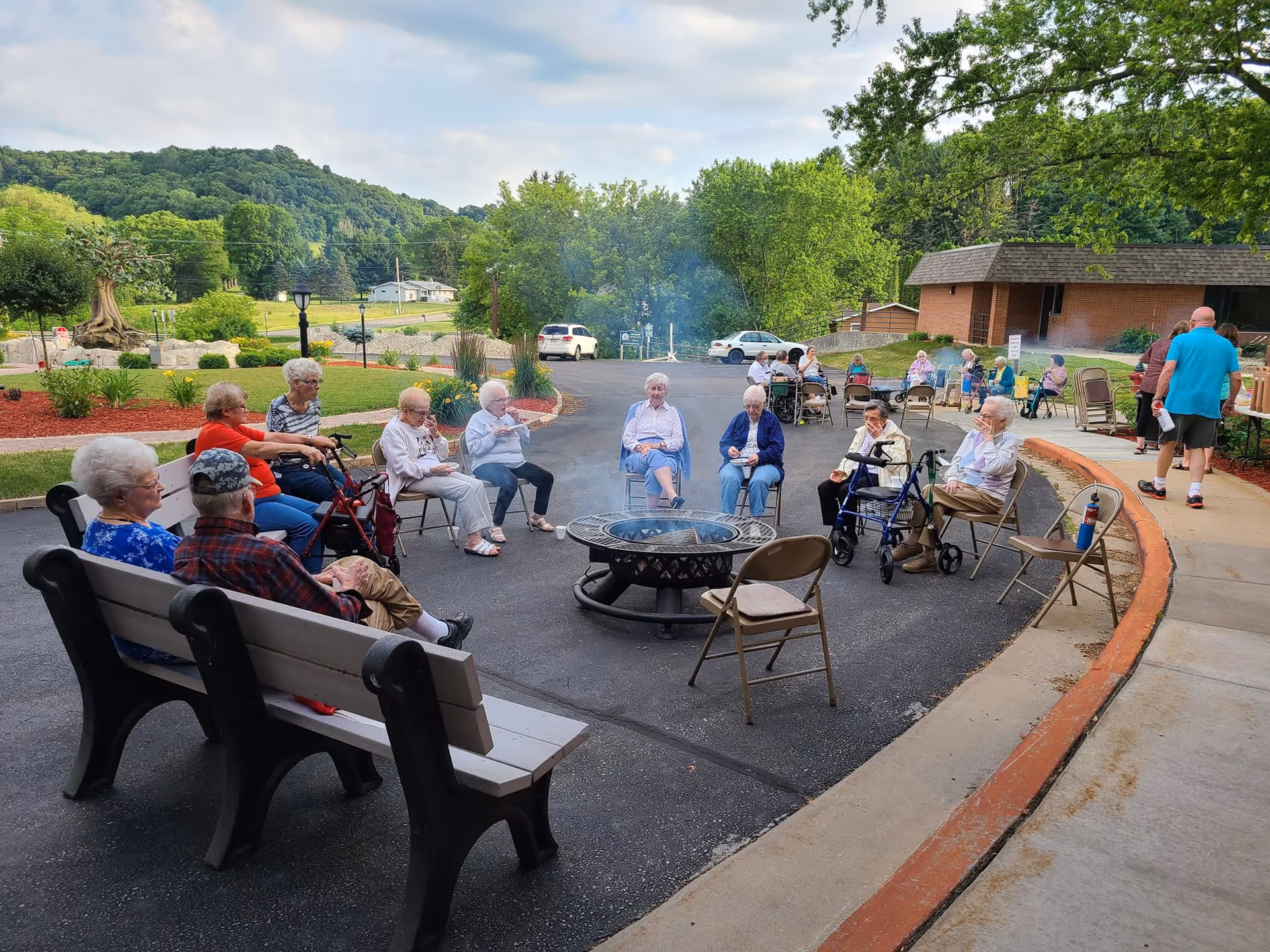 A group of elderly people sitting outdoors around a fire pit on chairs and benches, with some using walkers. They are in a paved area near a building with a brick exterior, surrounded by greenery and hills in the background. A few people are standing near a table on the right side.