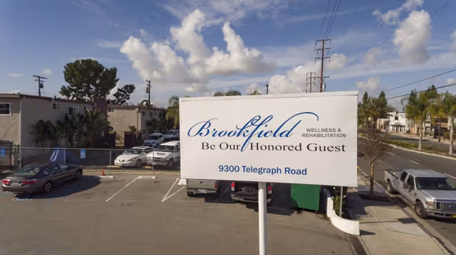 Outdoor view of a parking lot with a large white sign that reads 'Brookfield Wellness & Rehabilitation Be Our Honored Guest 9300 Telegraph Road'. Several cars are parked in the lot and residential buildings and trees are visible in the background under a partly cloudy sky.