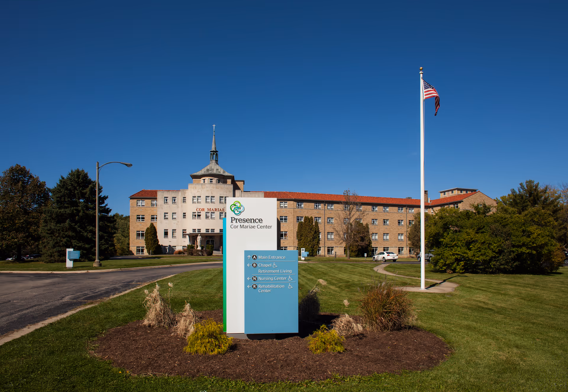 Exterior view of the Ascension Living Cor Mariae Village building with a clear blue sky. In front of the building is a sign for Presence Cor Mariae Center with directions to the main entrance, chapel, retirement living, nursing center, and rehabilitation center. There is a flagpole with an American flag on the right side and well-maintained grass and landscaping around the area.