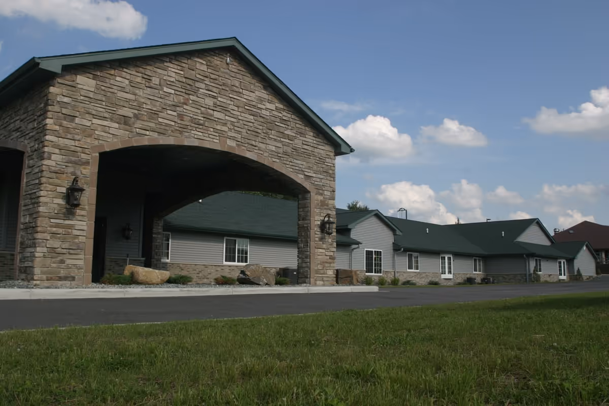 Exterior view of Acorn Hill Apartments showing a long, single-story building with a stone archway entrance, gray siding, green roof, and a paved driveway with grass in the foreground under a partly cloudy sky.