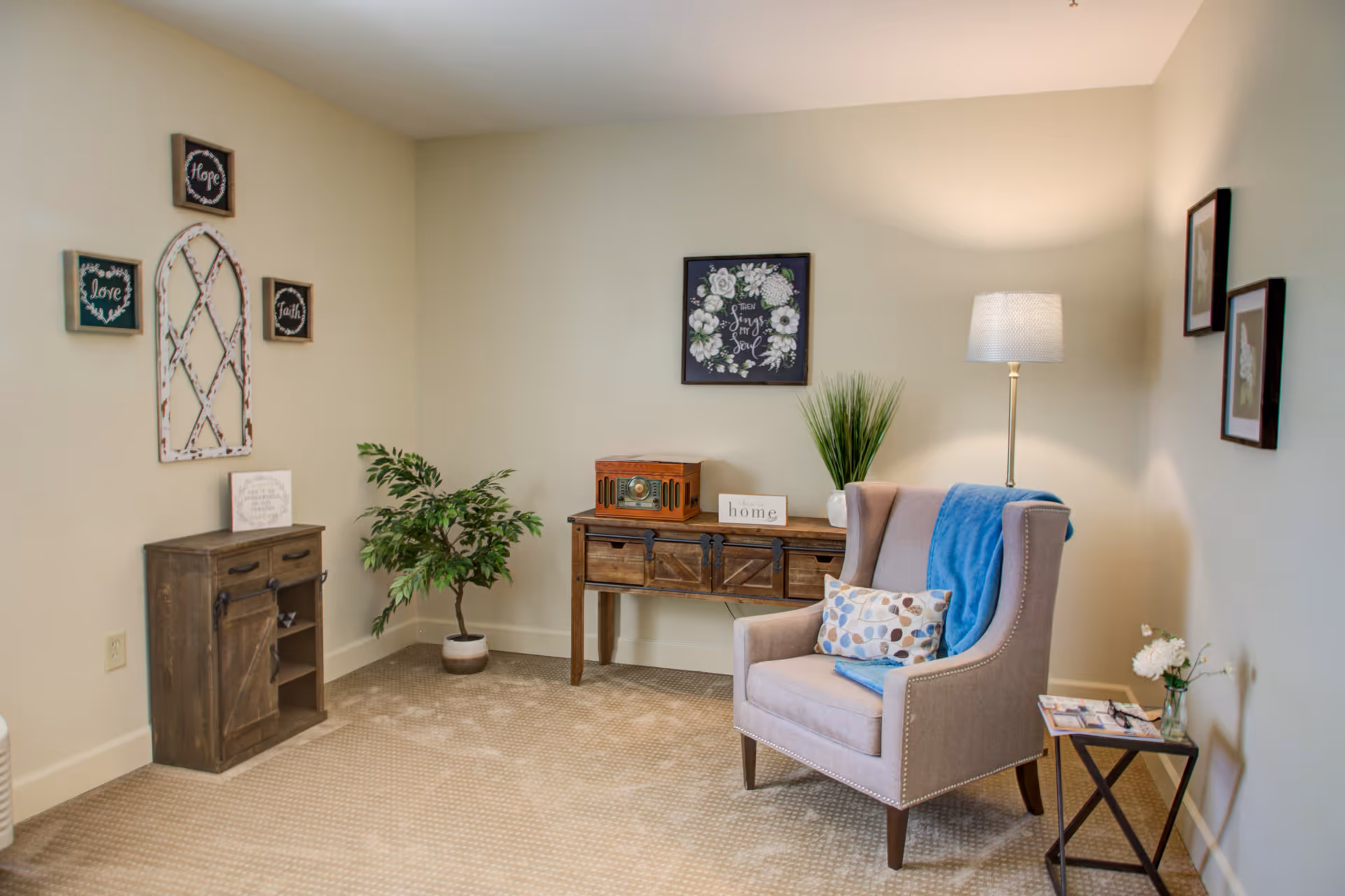 A cozy living room corner with a beige armchair draped with a blue throw and a patterned pillow. Next to the chair is a small side table with a vase of white flowers and magazines. Behind the chair is a floor lamp providing warm light. A wooden console table against the wall holds a vintage radio, a small plant, and a decorative sign that says 'home'. On the left side, there is a small wooden cabinet and a potted plant. The walls are decorated with framed artwork and inspirational signs.