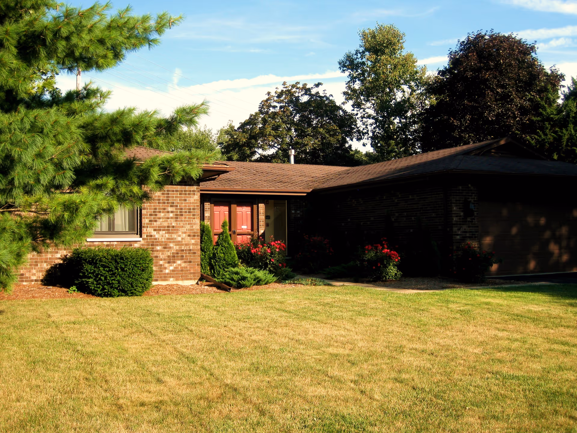 A single-story brick house with a brown roof surrounded by green trees and bushes. The front yard has a well-maintained lawn and some flowering plants near the entrance. The sky is clear with some clouds.