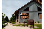 Exterior view of a gray wooden building with a covered porch and a brown bench on a concrete pathway surrounded by grass and landscaping under a cloudy sky.