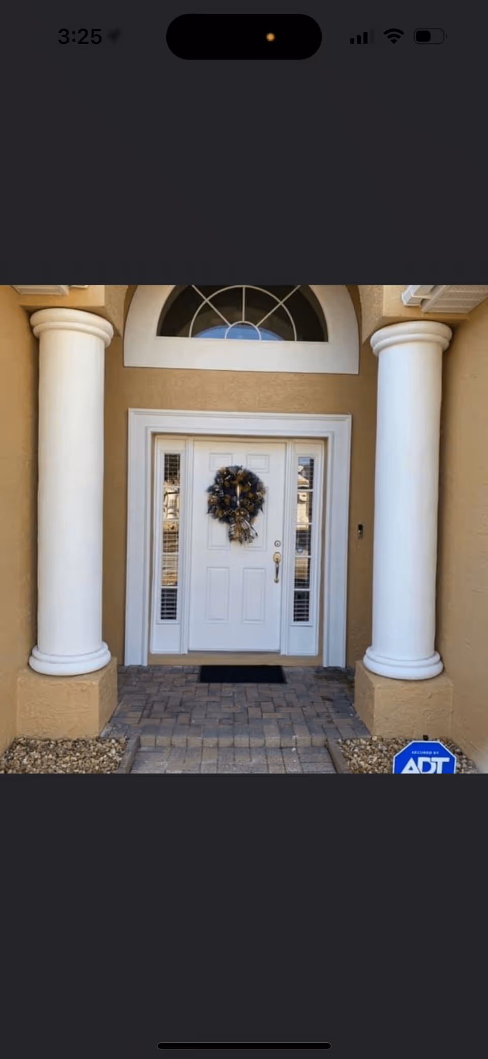 Front entrance of a building with a white door decorated with a dark-colored wreath. The entrance is flanked by two large white columns and has a semi-circular window above the door. The ground is paved with bricks, and there is an ADT security sign on the right side near the base of the column.