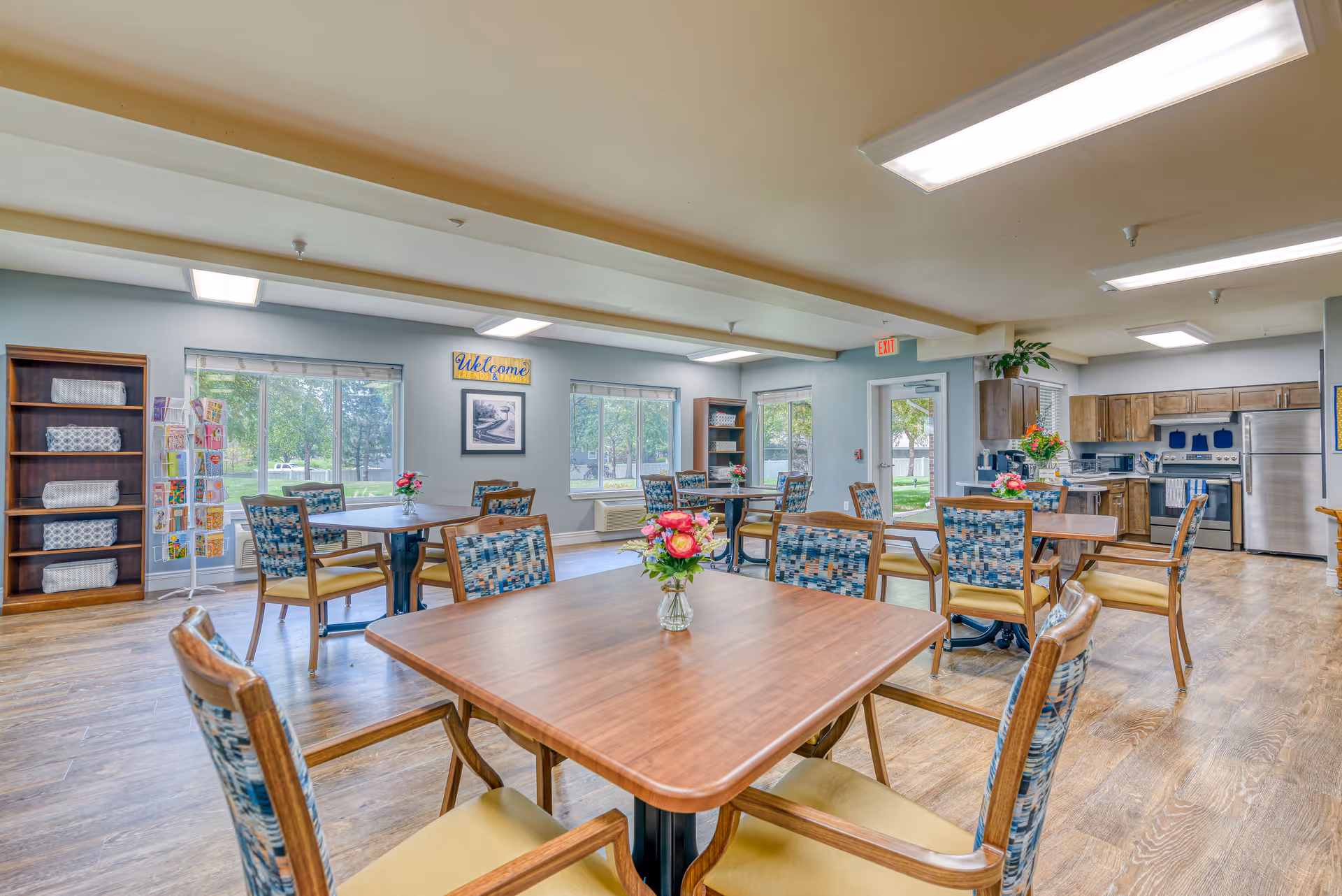 A bright and spacious dining area in a senior living facility with several wooden tables and chairs arranged neatly. Each table has a small vase with colorful flowers. Large windows let in natural light and provide a view of green trees outside. The room features wooden flooring, a kitchen area with stainless steel appliances, and shelves with folded linens and greeting cards. A 'Welcome' sign is displayed on the wall.