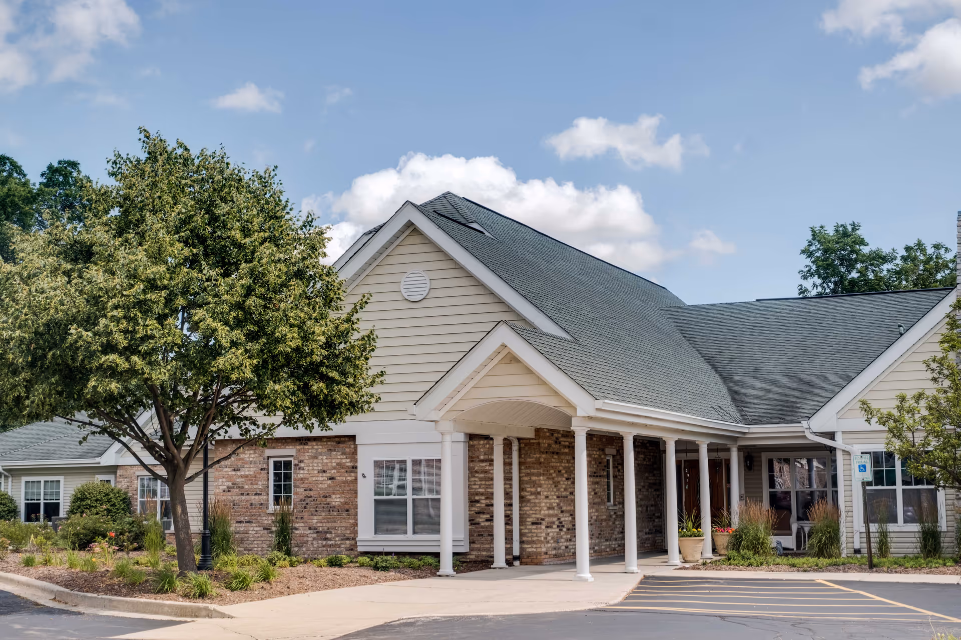 Exterior view of a single-story assisted living facility building with beige siding and brick accents. The building has a covered entrance supported by white columns, surrounded by landscaped greenery and trees under a partly cloudy sky.