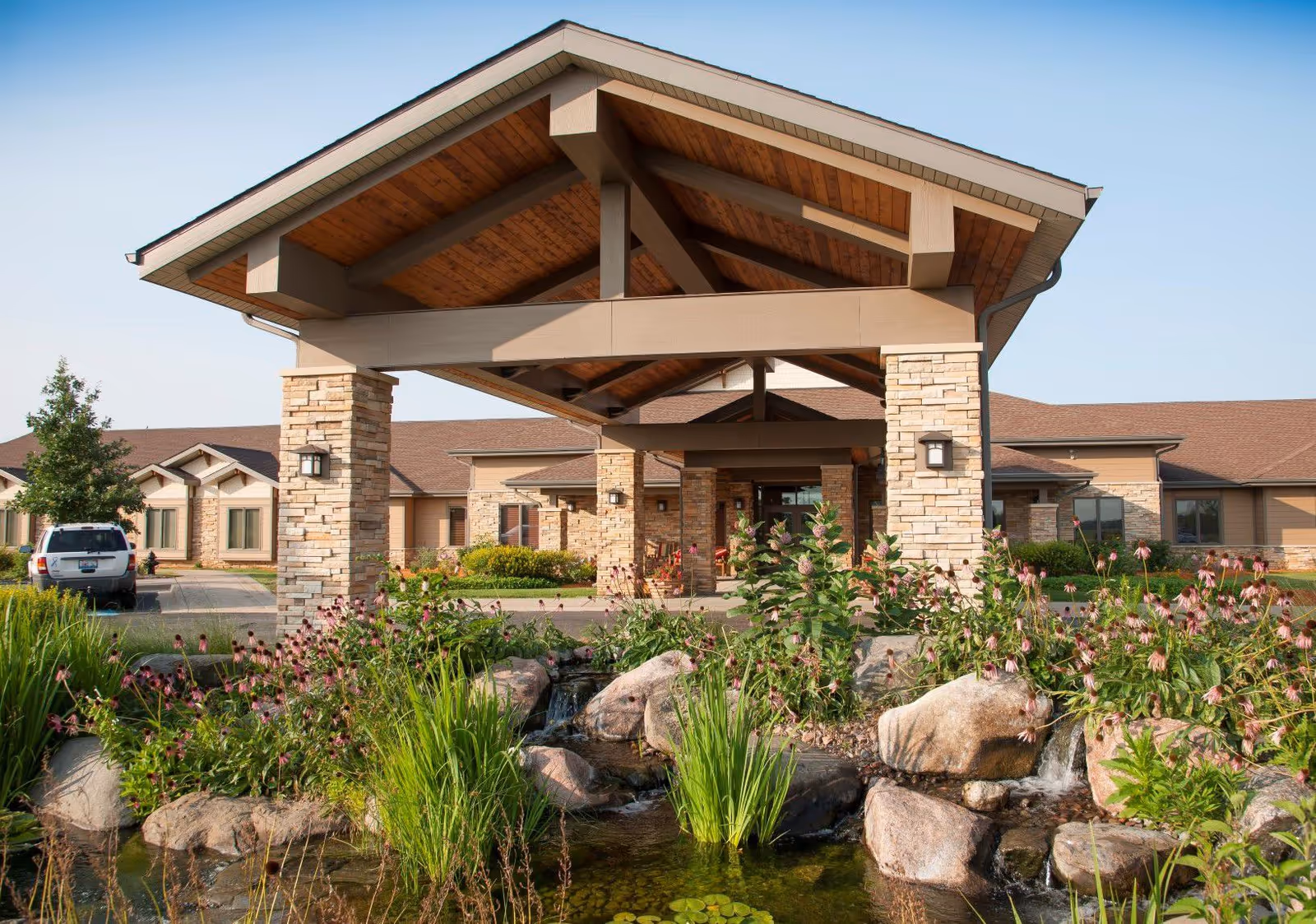 Entrance of NorthPointe Terrace facility featuring a covered driveway with stone pillars and a wooden roof. In the foreground, there is a landscaped garden with rocks, green plants, and flowers surrounding a small water feature. The building has a beige exterior with multiple windows and a brown roof.