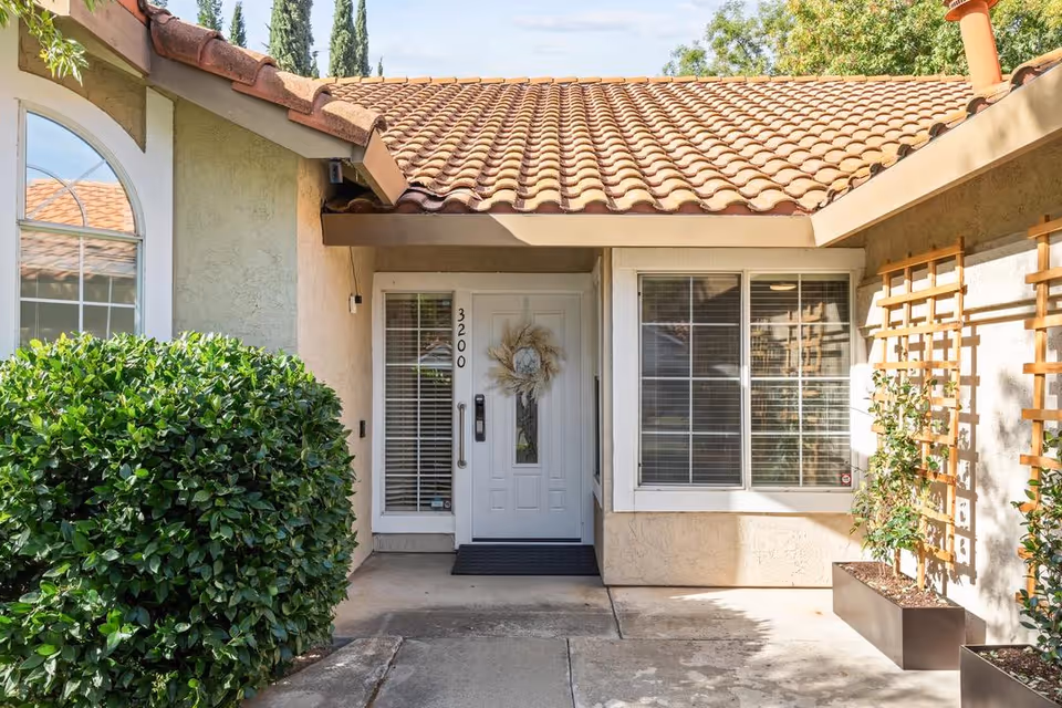 Entrance to a residential unit with a white door decorated with a wreath, flanked by a window with blinds and a glass panel with the number 3200. The exterior features beige stucco walls, a tiled roof, green bushes, and potted plants on a concrete walkway.