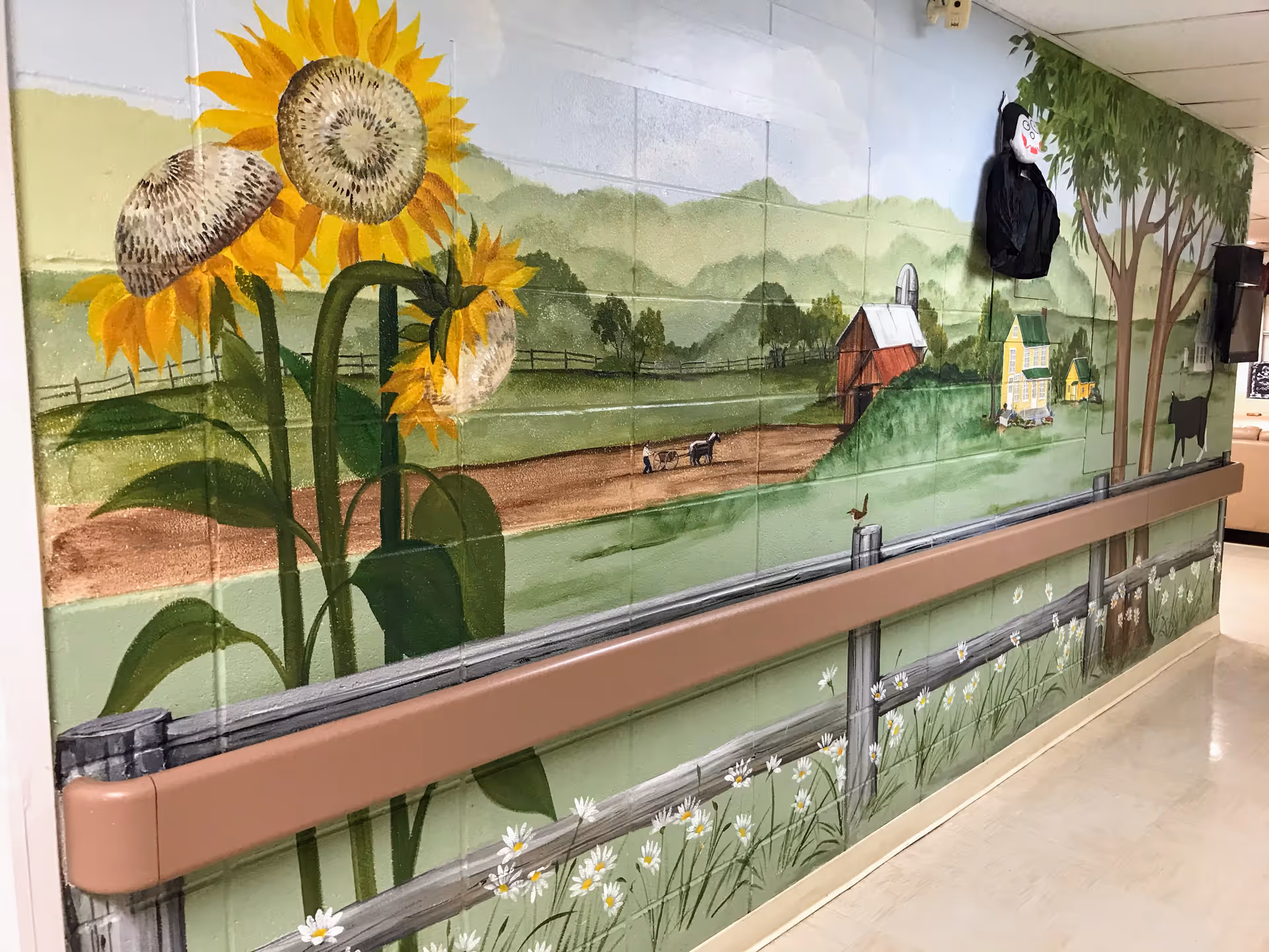 A mural painted on a hallway wall depicting a rural farm scene with large sunflowers, a wooden fence, a dirt road with a horse and cart, a red barn, a yellow farmhouse, trees, and a cow. The hallway has a handrail along the wall and a light-colored floor.