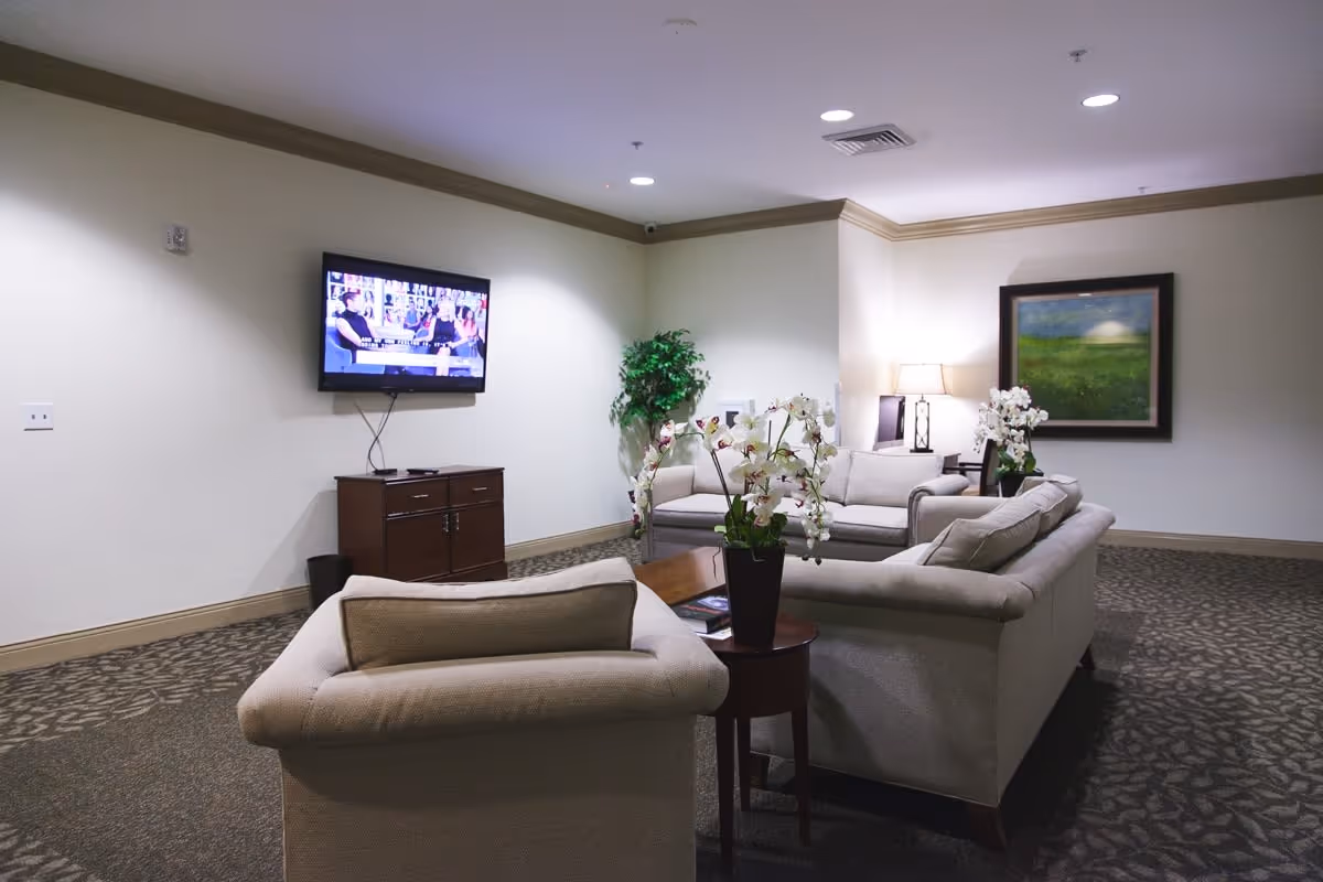 A cozy senior living common area with beige sofas arranged around a wooden coffee table. There are potted white orchids on the table and side table. A flat-screen TV is mounted on the wall above a wooden cabinet. A green potted plant is in the corner, and a framed landscape painting hangs on the wall. The room is softly lit with ceiling lights and a table lamp.