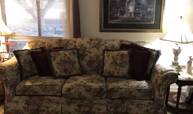 A floral patterned sofa with multiple cushions in a living room setting. Behind the sofa is a window with blinds partially open, letting in natural light. On the right side, there is a side table with a lamp and decorative items, and a framed picture hanging on the wall above the sofa.