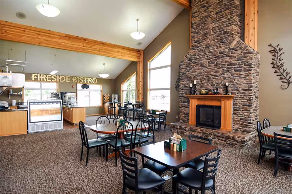 Interior view of a dining area named Fireside Bistro with multiple tables and chairs arranged on a carpeted floor. A large stone fireplace with a wooden mantle is on the right side, and large windows allow natural light to fill the room. The ceiling has wooden beams and hanging light fixtures.