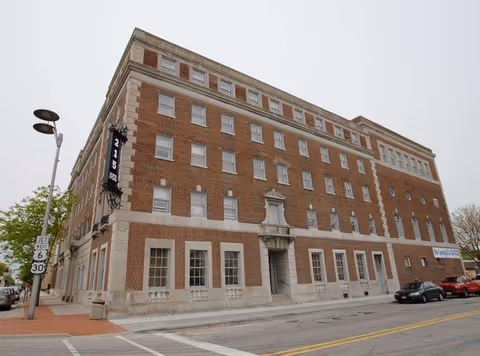 Exterior view of a large multi-story brick building with numerous windows and a decorative entrance, located at a street corner with a traffic sign indicating junction of routes 6 and 30.