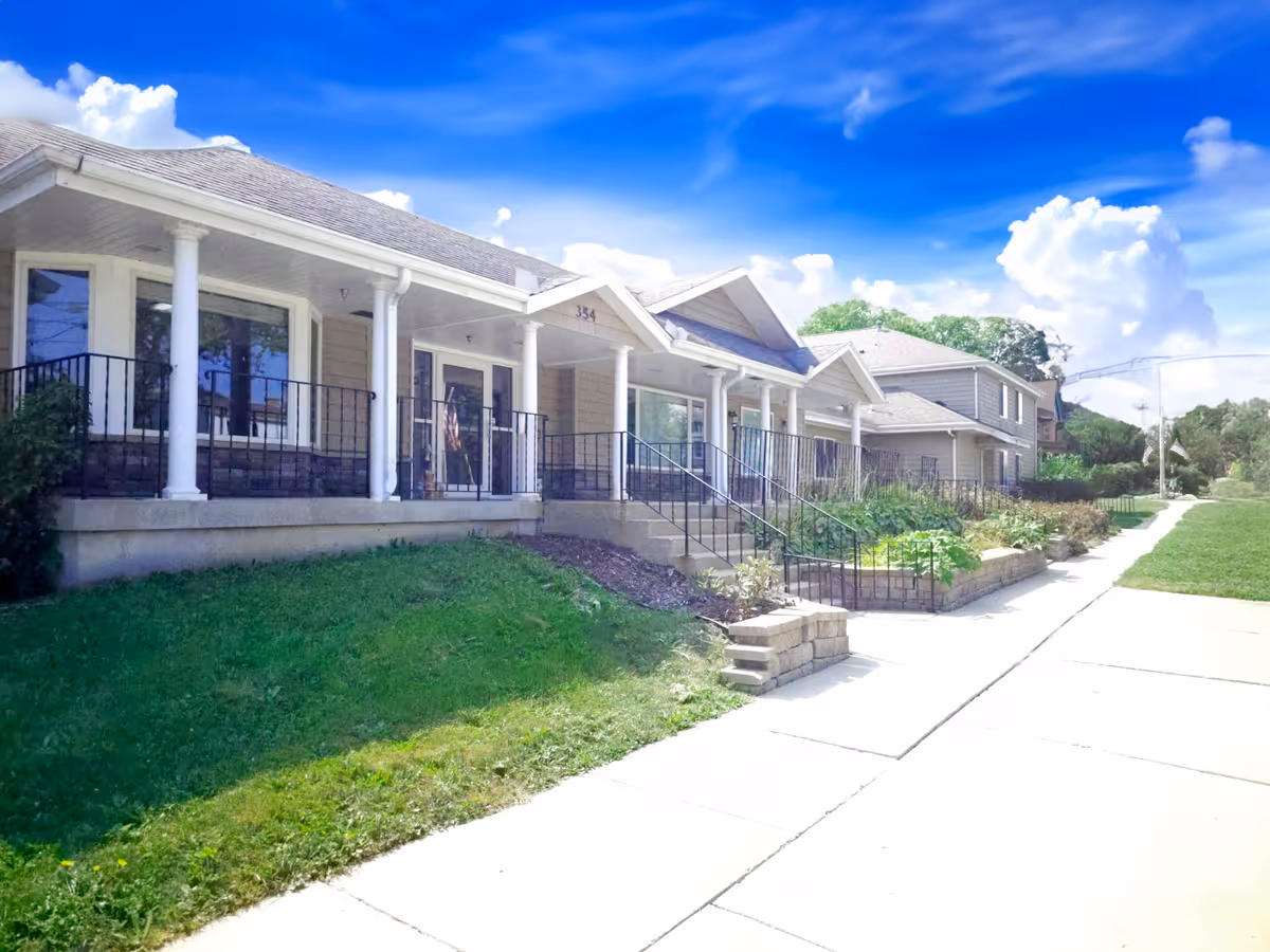 Exterior view of a senior living facility building with a covered porch supported by white columns, black railings, and steps leading up to the entrance. The building is surrounded by green grass, plants, and a sidewalk under a bright blue sky with scattered clouds.
