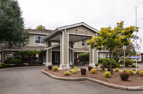 Exterior view of Brookdale River Road senior living facility entrance with a covered drop-off area, surrounded by landscaped bushes and trees.
