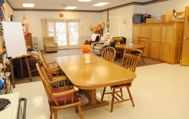 A cozy common area in Concordia Haven Apartments featuring a wooden dining table with six chairs, a whiteboard on an easel, a sofa and armchairs near the back wall, and wooden cabinets along the right side. The room has light-colored walls and flooring, with a glass door leading outside.