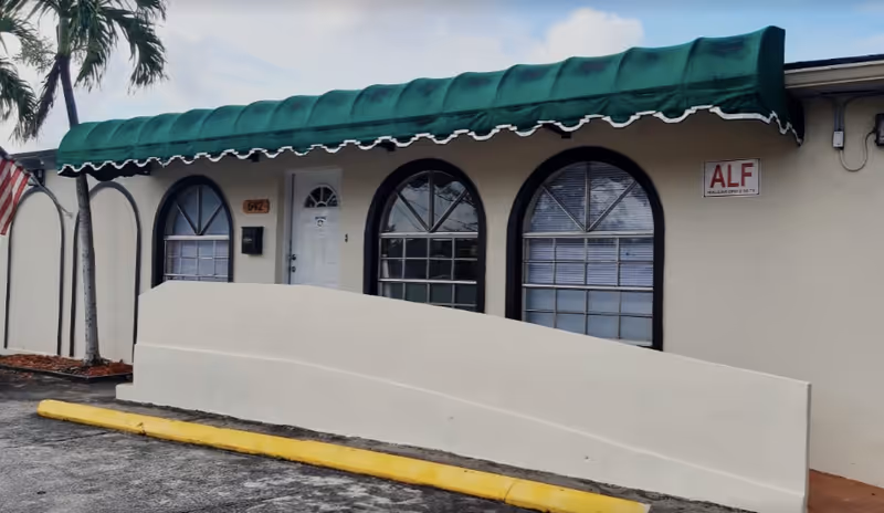 Exterior view of a single-story building with a green awning above three arched windows and a white door. There is a small palm tree on the left side and a yellow curb in front of the building. A sign on the wall reads 'ALF'.