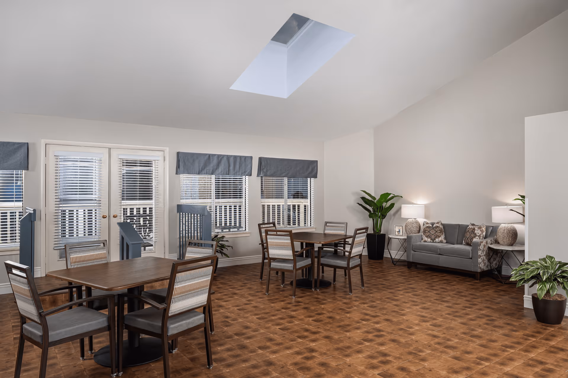 A spacious senior living common area with two wooden tables each surrounded by four chairs. There is a gray sofa with patterned cushions against the wall, flanked by two side tables with lamps. Several potted plants add greenery to the room. The floor is a warm-toned wood pattern, and the walls are painted light gray. A skylight in the ceiling allows natural light to brighten the space. French doors and windows with blinds and blue valances are visible on one side.