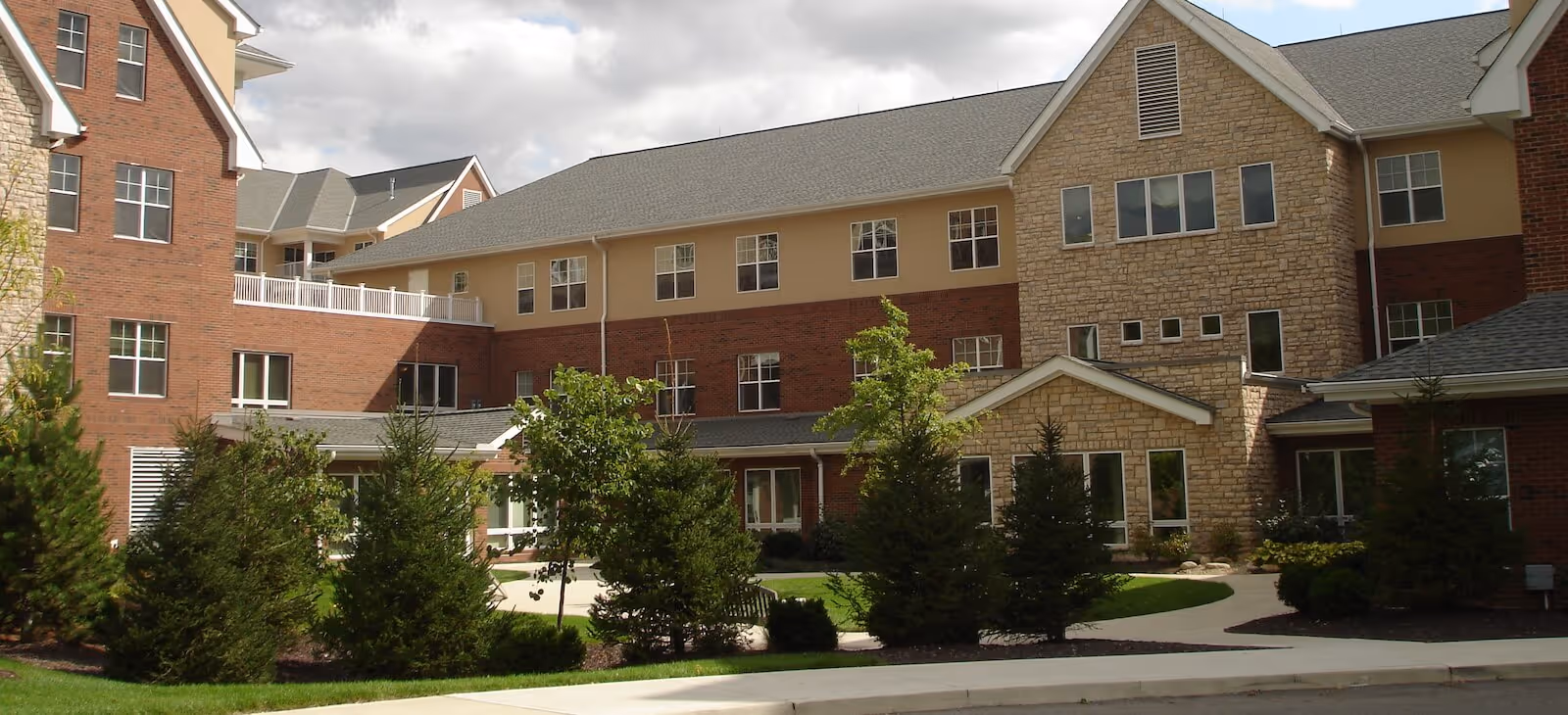 Exterior view of a senior living facility building with a mix of brick and stone facade, multiple windows, and a landscaped area with trees and shrubs in front under a partly cloudy sky.