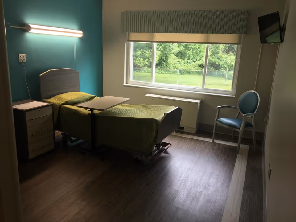 A small, simple nursing facility room with a single hospital bed covered with a green blanket, a bedside table with drawers, an adjustable overbed table, a teal upholstered chair, a wall-mounted television, and a large window showing greenery outside.